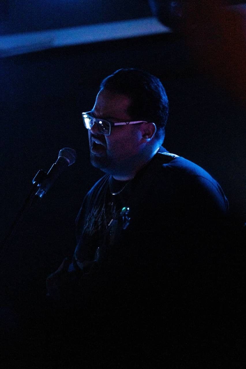 A man with short dark hair and glasses sings passionately into a microphone under blue stage lighting, creating a dramatic and moody atmosphere.