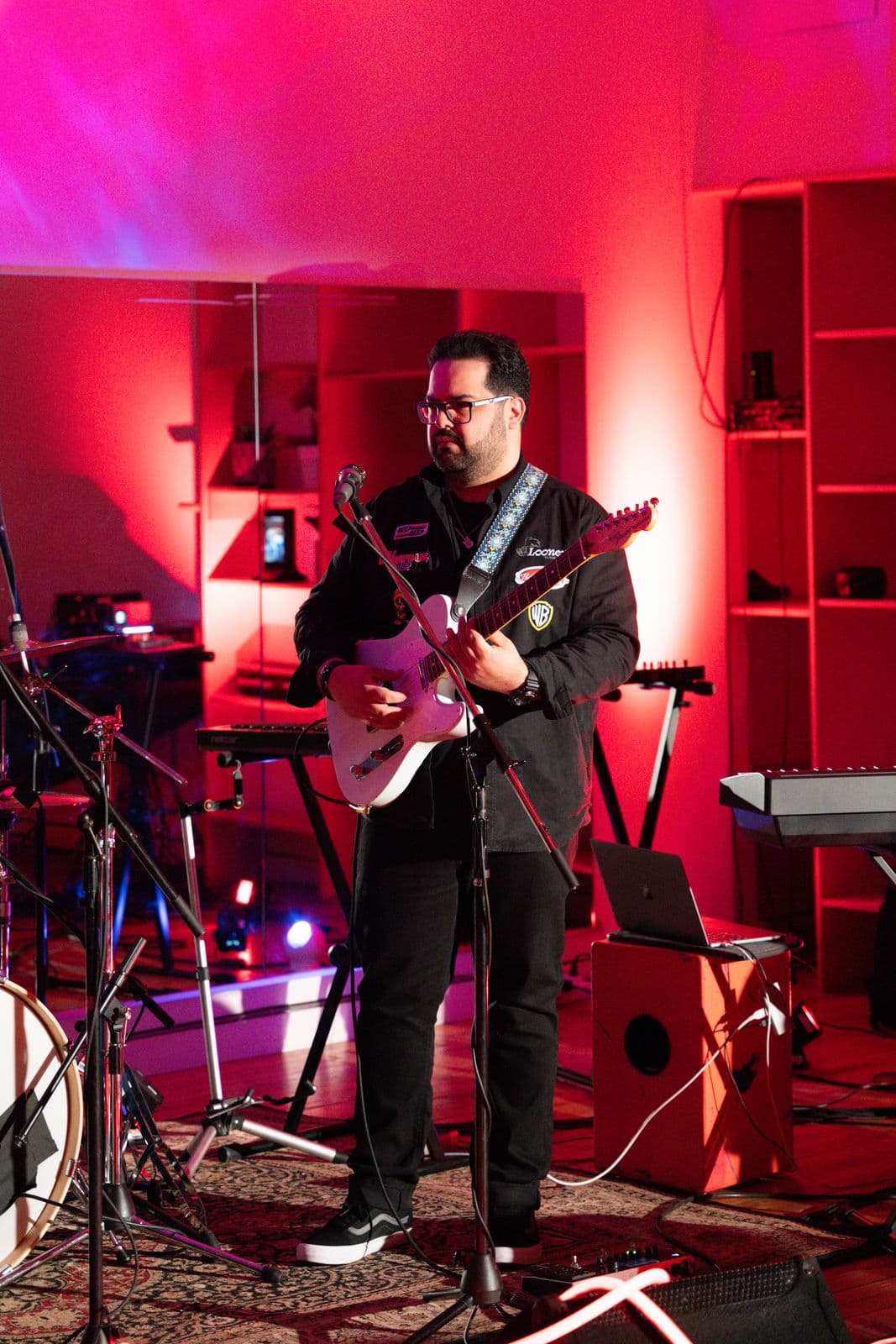 A guitarist with glasses and a black jacket plays an electric guitar on stage, surrounded by musical instruments and bathed in vibrant red lighting.