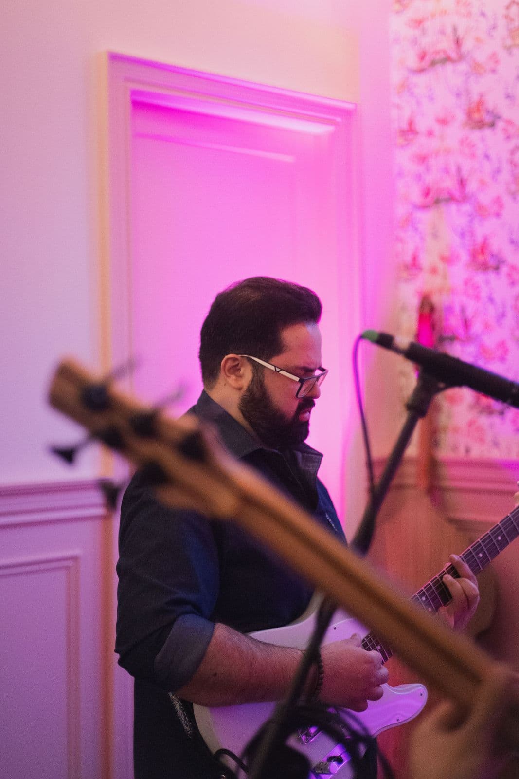Arash with glasses leading the house band at a Beytooteh show, deeply focused while playing electric guitar in a softly lit room with pink and floral accents.