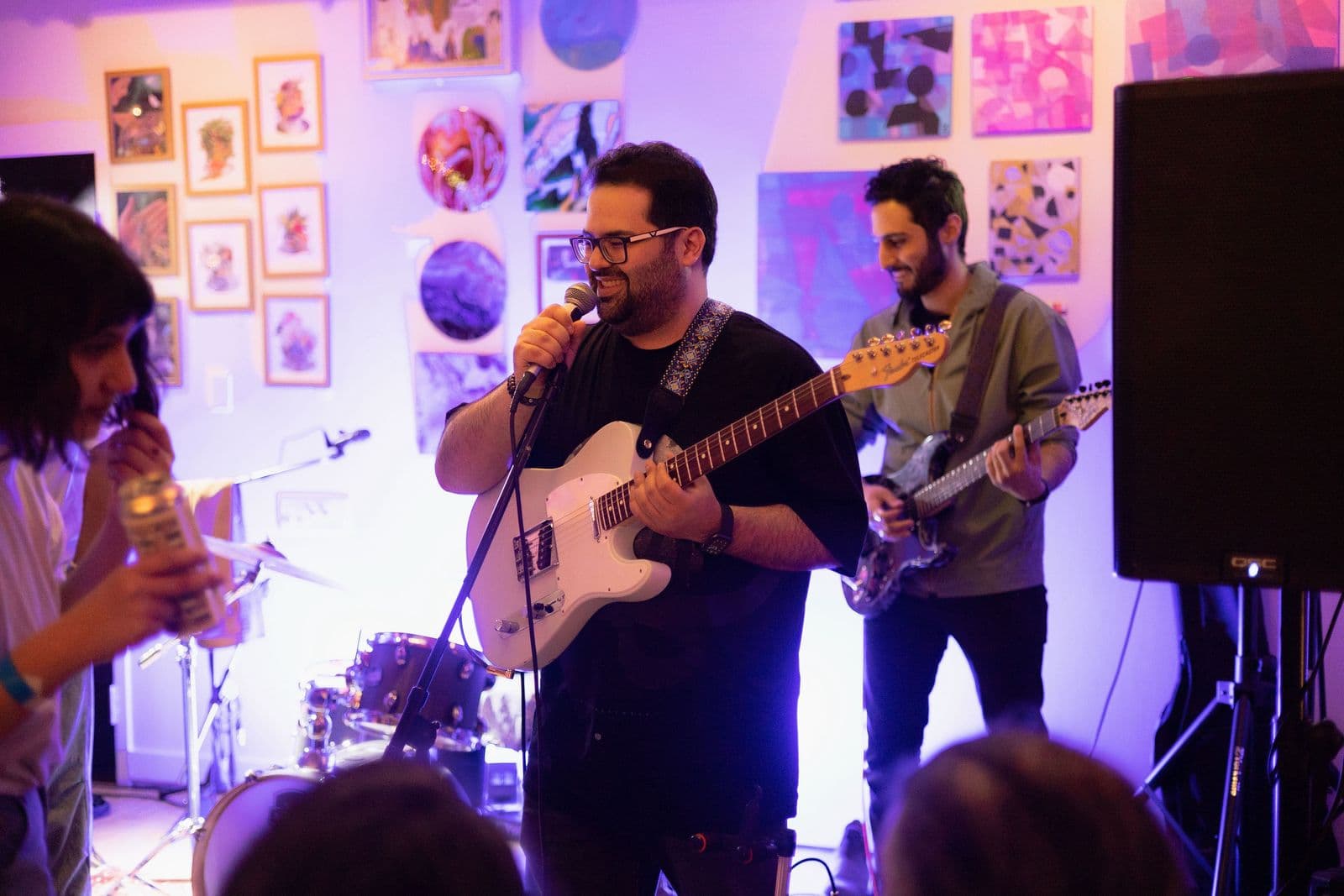 Edrock performing their song Toro Mikham live for the first time at a Beytooteh show — Arash singing into a microphone while another guitarist plays in the background of an art-filled room.
