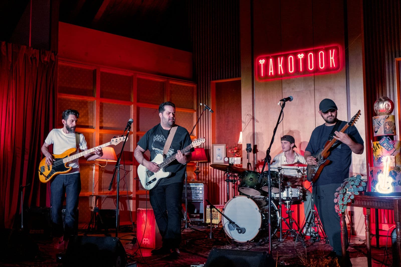 Four-member band performs on stage at a venue called TAKOTOOK, with a red neon sign, featuring two guitarists, a bassist, and a drummer in a lively, warmly lit environment.
