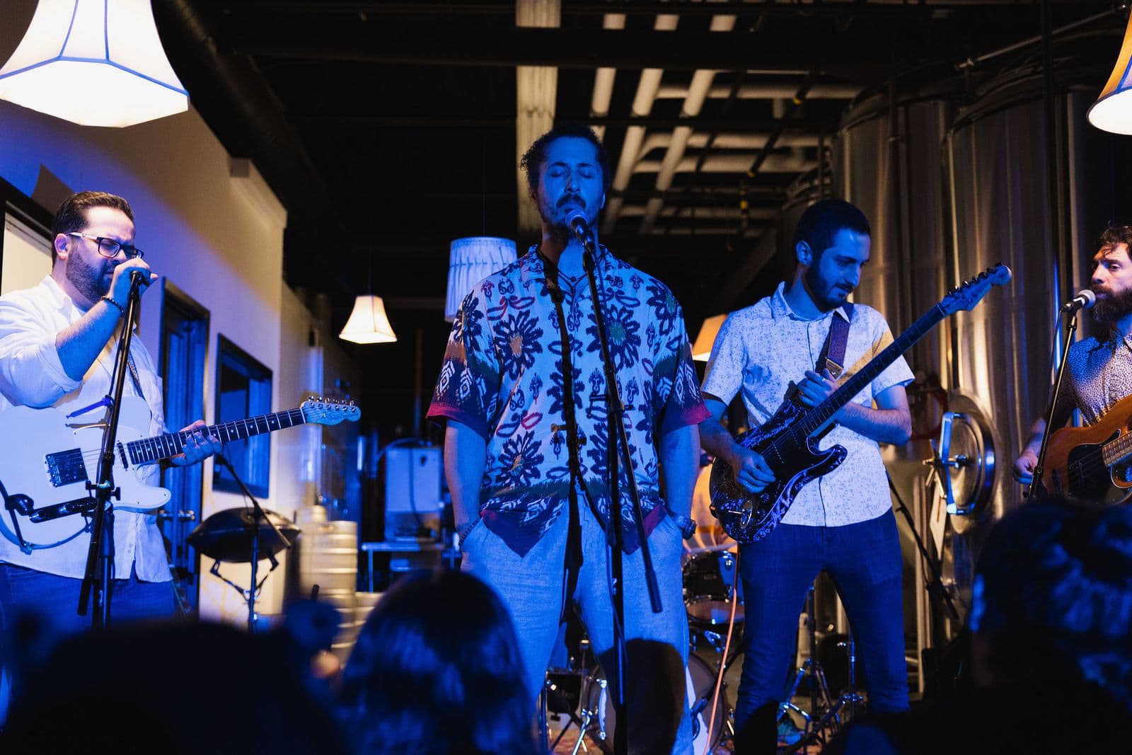Five-member band performs live under blue lights, featuring a lead singer in a patterned shirt, two guitarists, a bassist, and a drummer, with visible stage equipment and a crowd in the foreground.