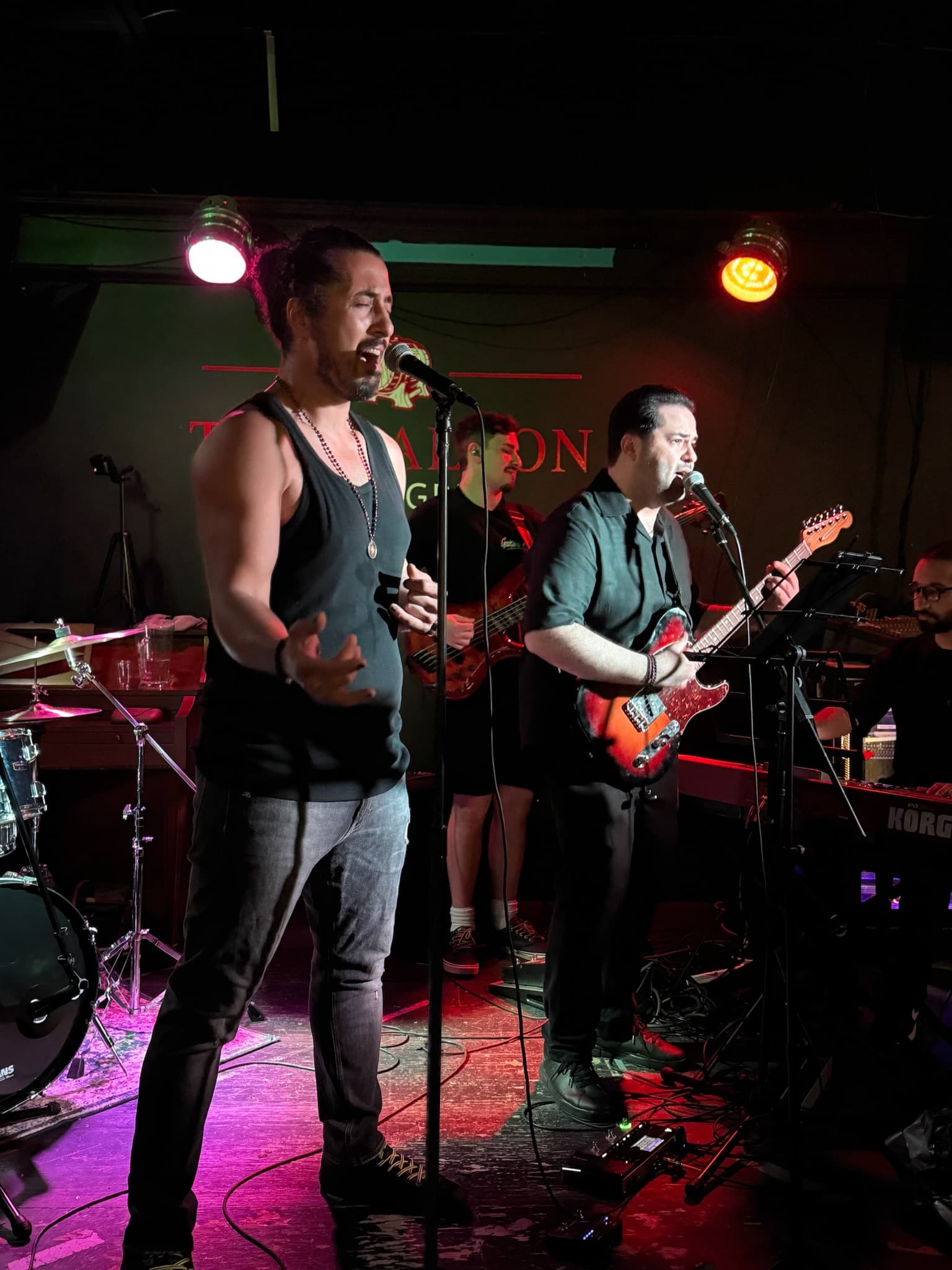 Two passionate vocalists perform on stage, one in a black tank top and the other in a black shirt playing guitar, with a backing band and colored spotlights creating an energetic mood.