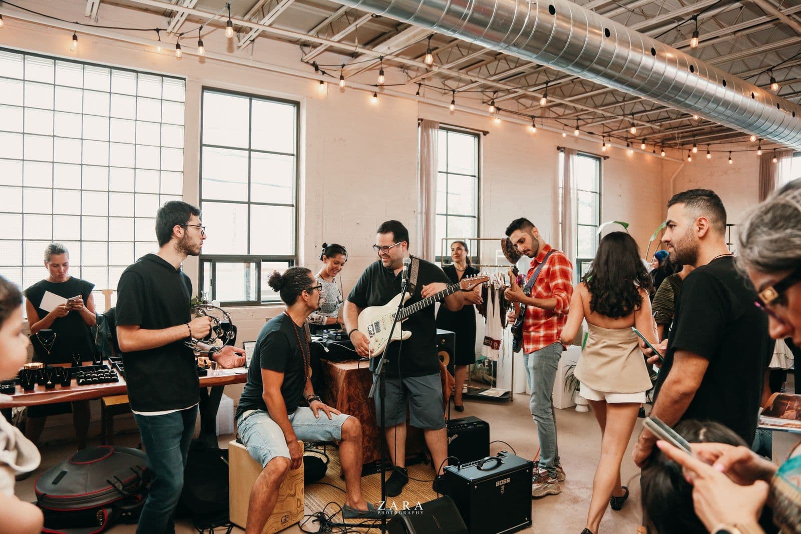 Indoor jam session with multiple musicians, including a guitarist, percussionist, another guitarist in plaid, and plenty of people enjoying the relaxed atmosphere in a bright, industrial space.