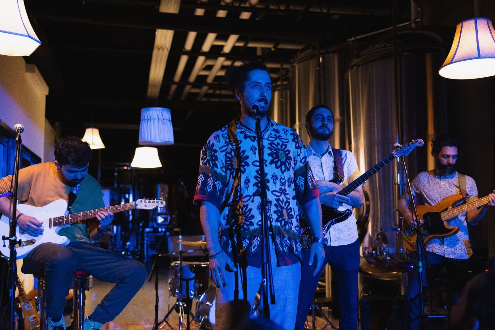 Four-member band performs live under blue ambient lighting, featuring a seated guitarist, a central vocalist in a vibrant patterned shirt, a lead guitarist, and a bassist, with metallic tanks and lamps in the background.