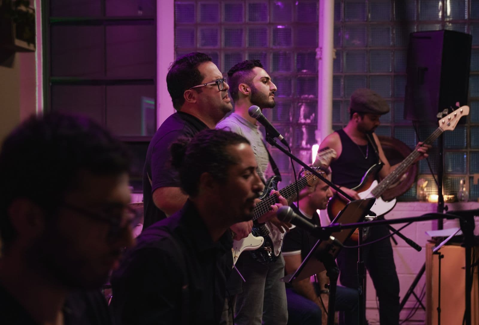 A band performs indoors under purple lighting, with the focus on two guitarists and other musicians playing keyboard and bass, all deeply engaged in their music.