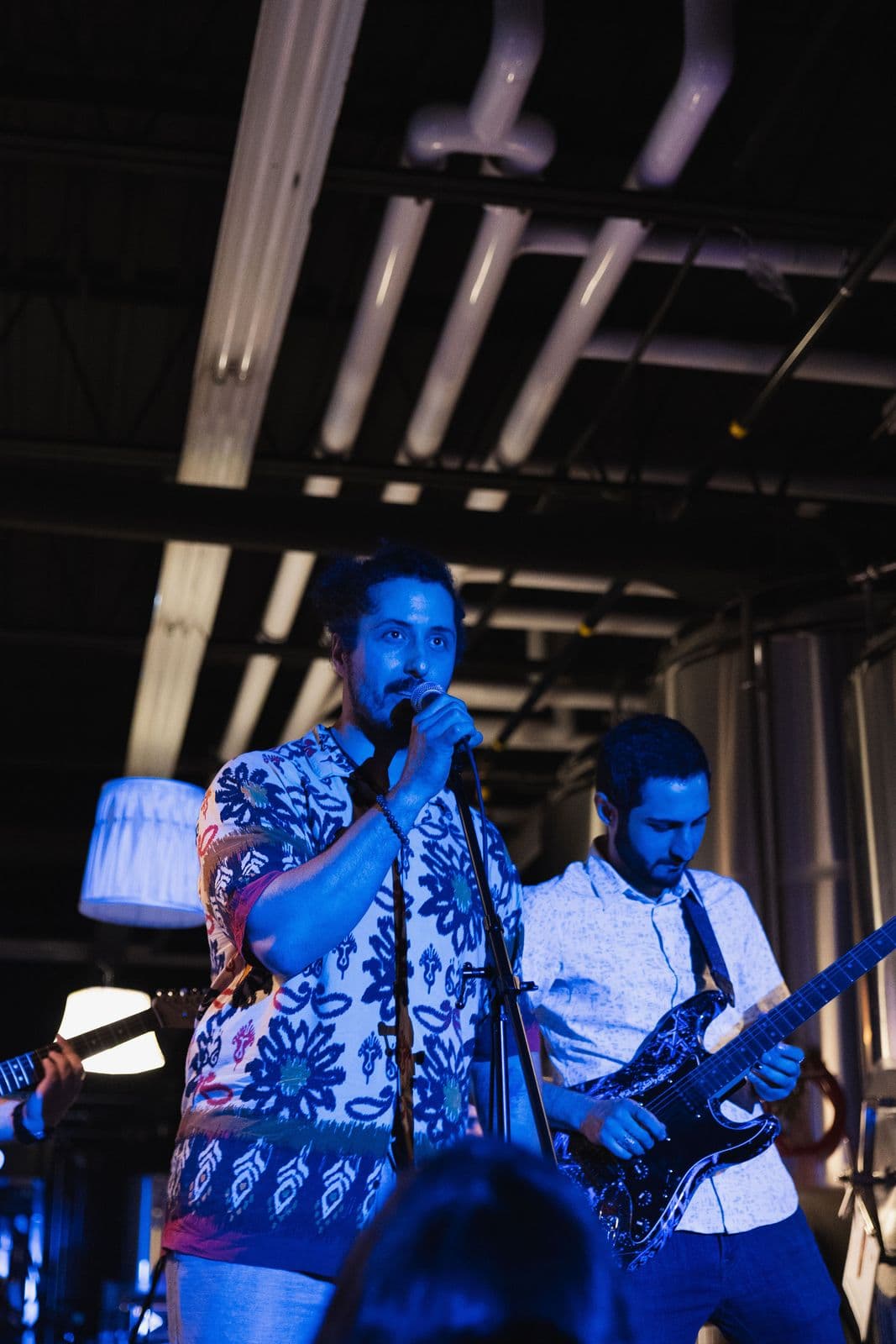 Close-up of a lead singer in a patterned shirt and a guitarist performing under blue lights, with industrial pipes and metal tanks in the background.