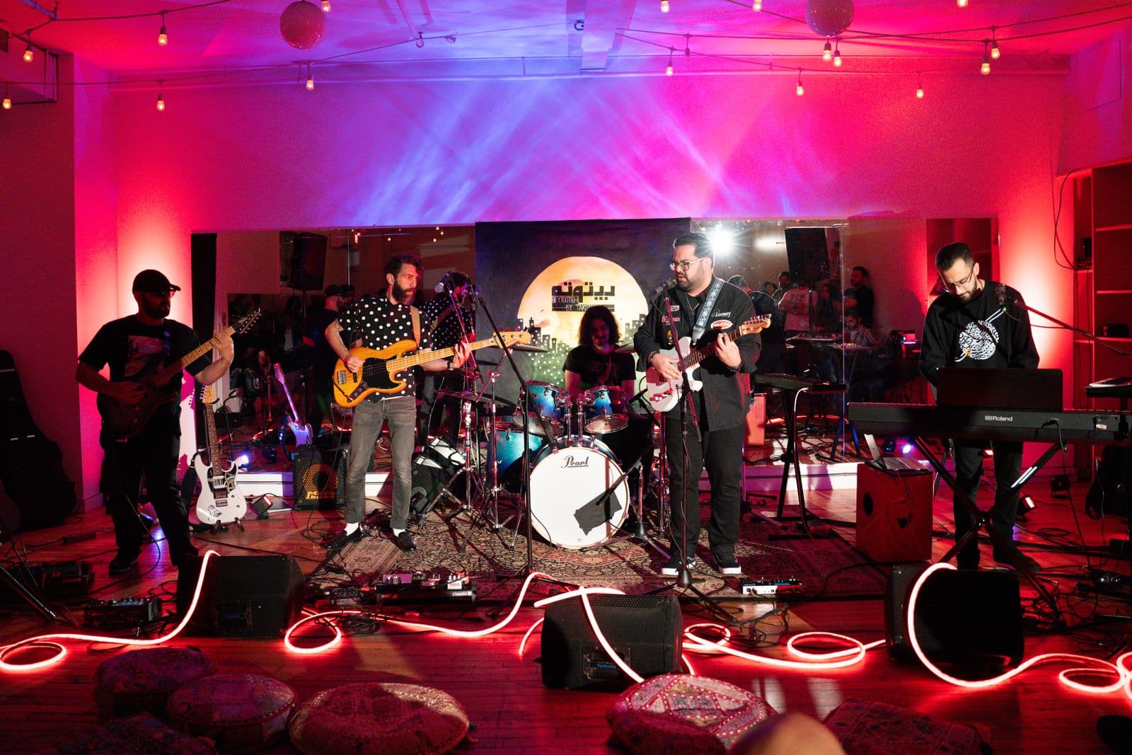 A full band performs onstage in a brightly lit studio, with five musicians playing guitar, bass, keyboards, and drums, surrounded by colorful lighting, mirrors, and decorative seat cushions.