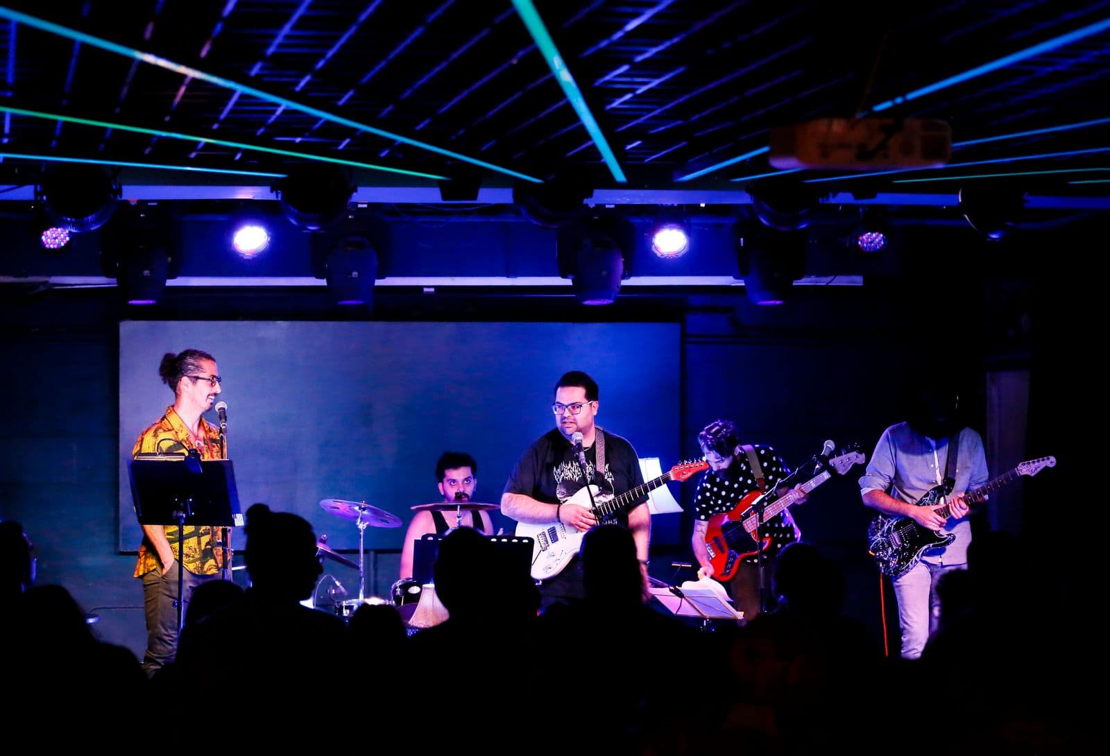 A five-member band performing on stage under vibrant blue and purple lighting, with audience silhouettes in the foreground and colorful ceiling lines above.