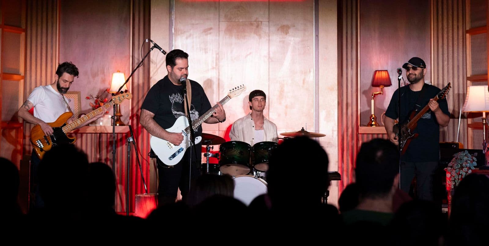Four-member band performs for a seated audience in a warmly lit venue with classic decor, featuring a lead guitarist and vocalist, bassist, drummer, and a second guitarist.
