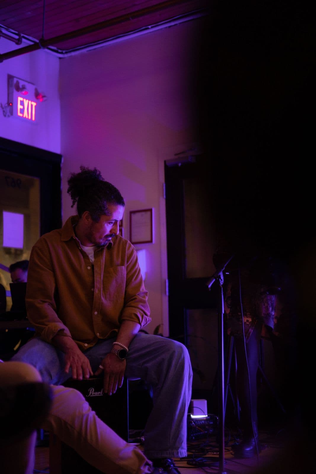 Musician with curly hair and a beard, wearing a brown shirt, sits on a cajón (box drum) under soft purple illumination, looking down thoughtfully during a performance.