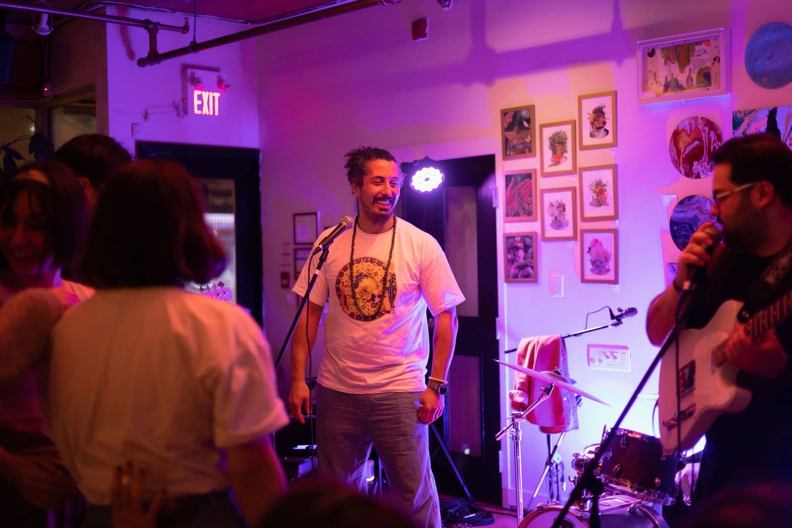 A smiling man in a white t-shirt stands at a microphone on stage, surrounded by audience members and musicians, bathed in soft purple lighting and ambient art decor.