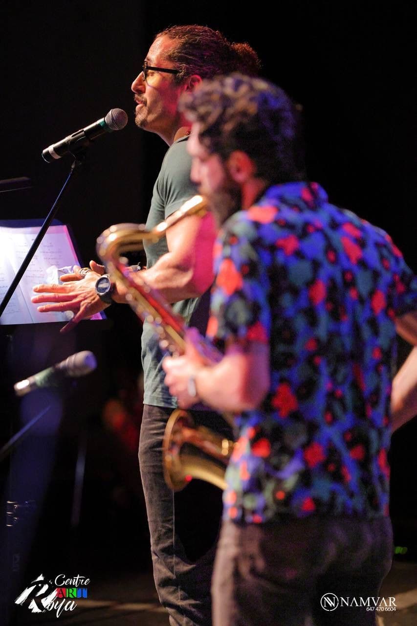 Two musicians perform on stage; the main focus is on the singer with glasses and tied-back hair speaking into a microphone while another plays the saxophone, both lit by dramatic, colorful stage lighting.