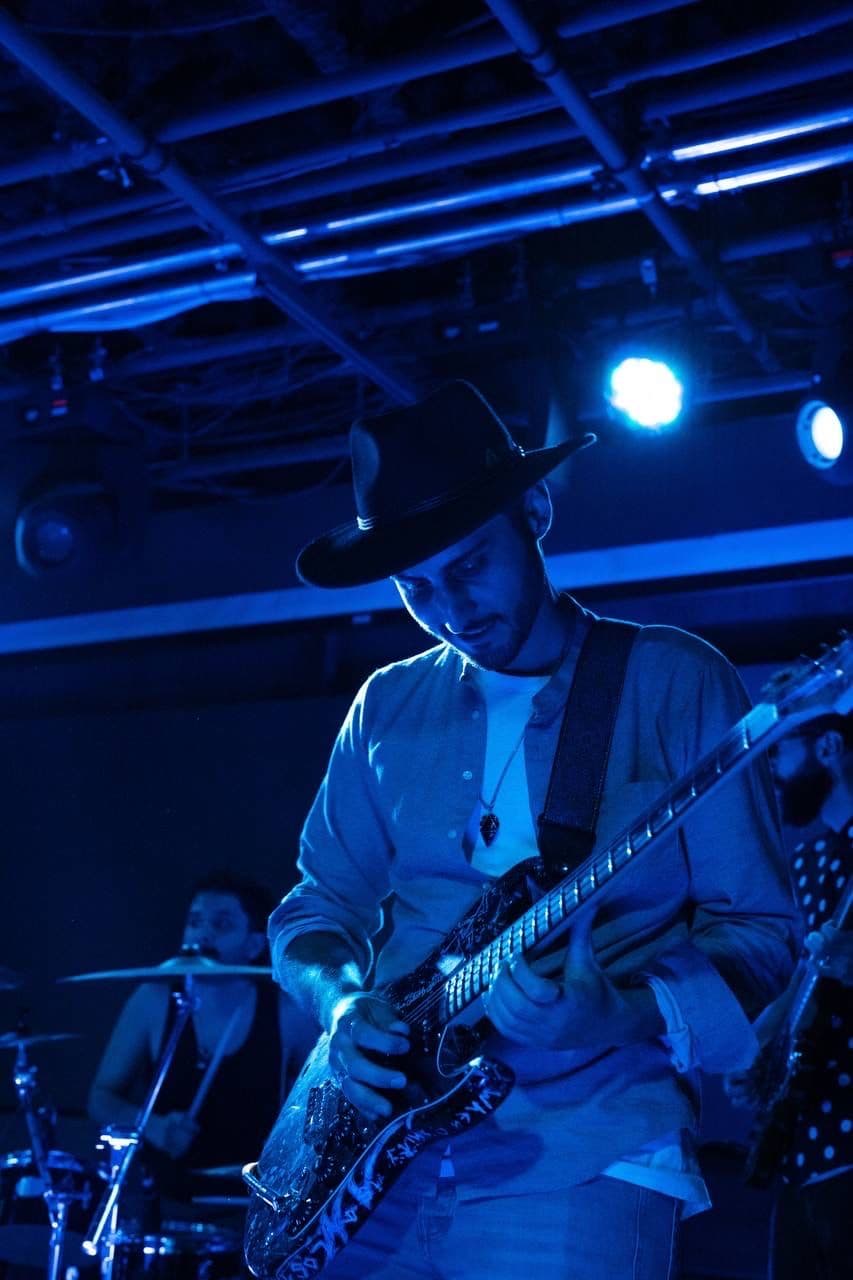 Guitarist wearing a wide-brim hat plays electric guitar intensely on stage under blue lighting, with a drummer visible in the background.
