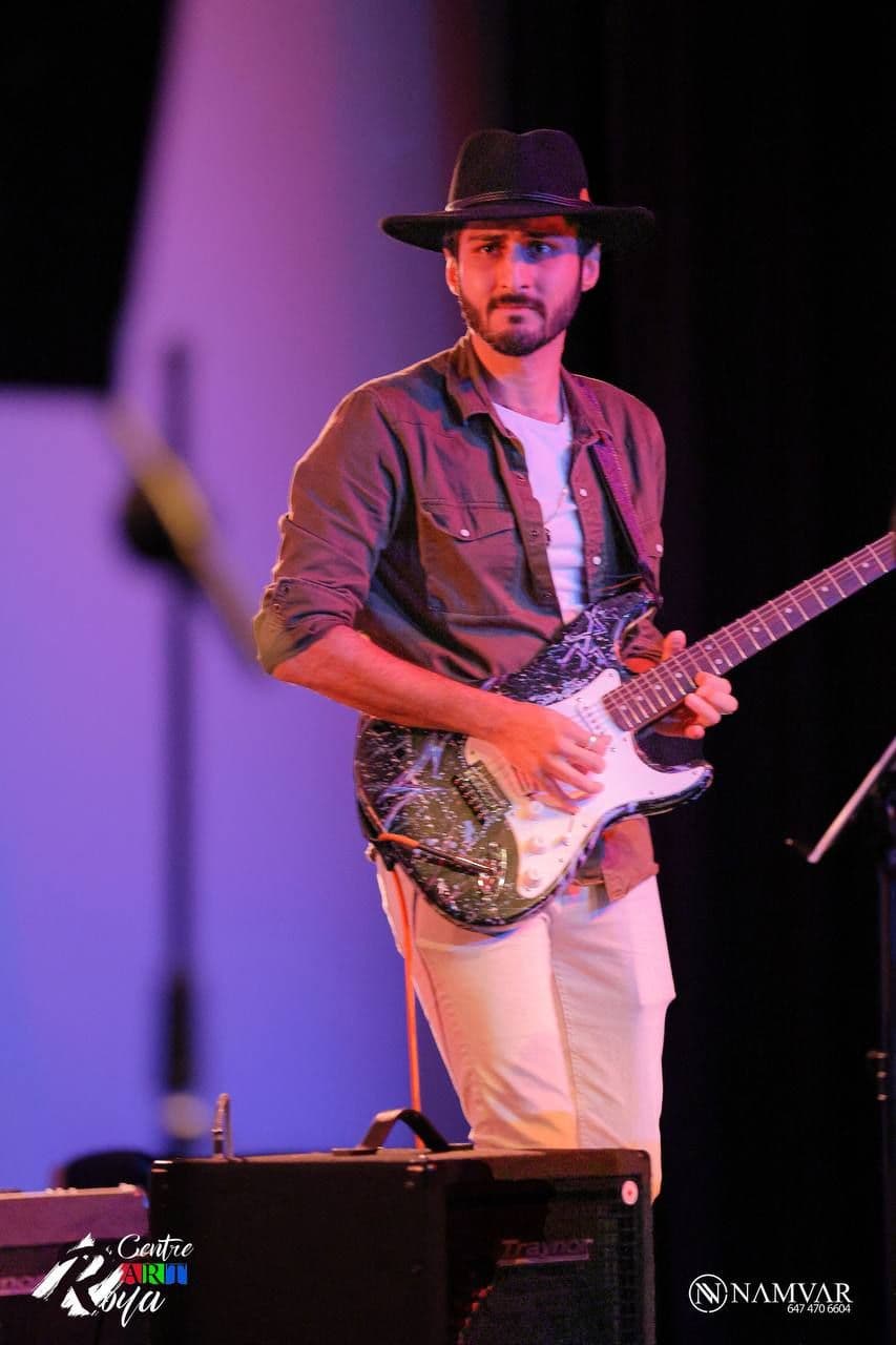 A focused guitarist in a hat and olive green jacket plays an electric guitar on stage at an indoor concert, standing in front of black curtain and amplifiers.