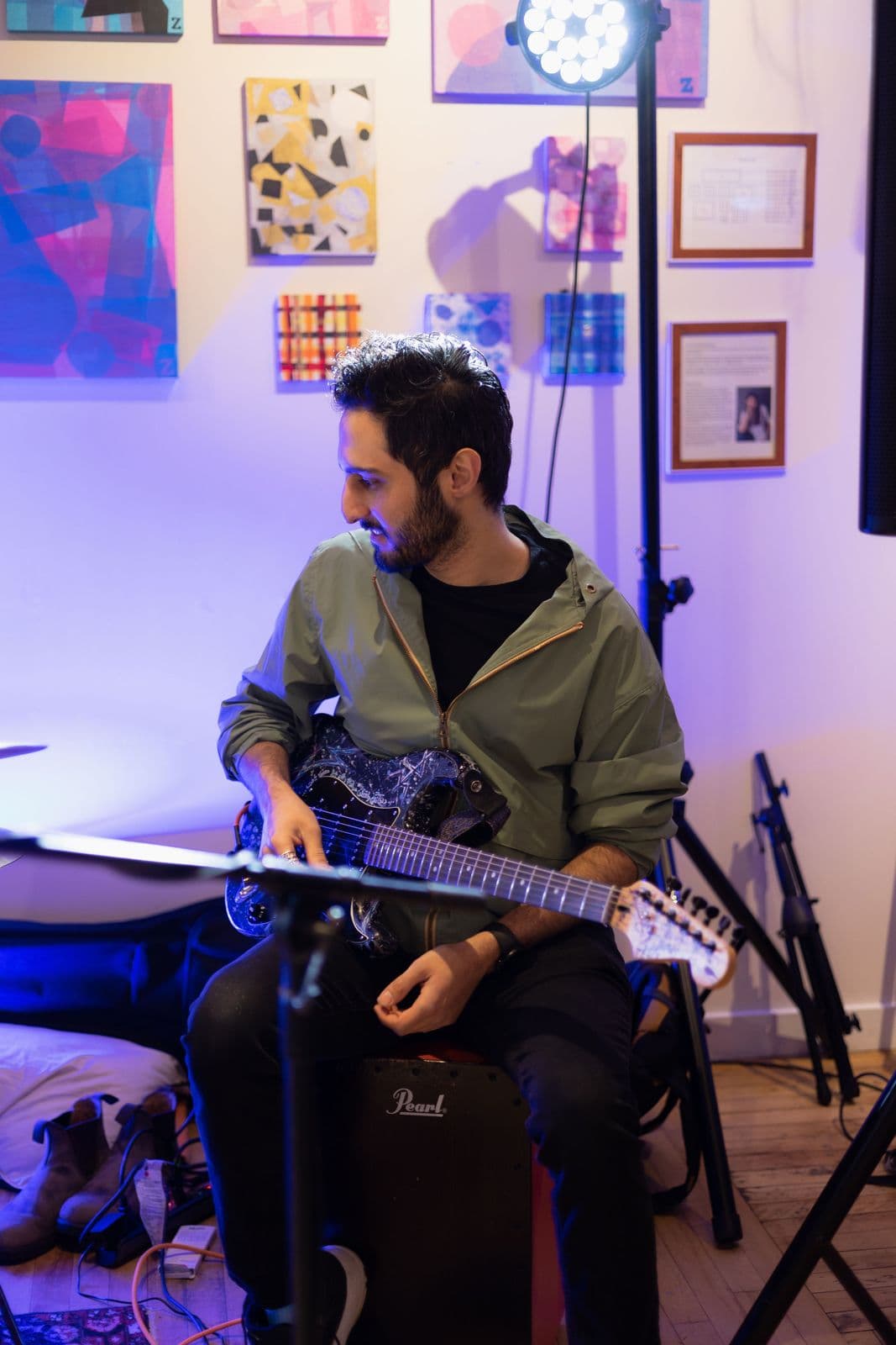 Guitarist in a light green jacket sits on a cajón with an electric guitar, surrounded by colorful abstract art and framed photos, illuminated by a bright stage light.
